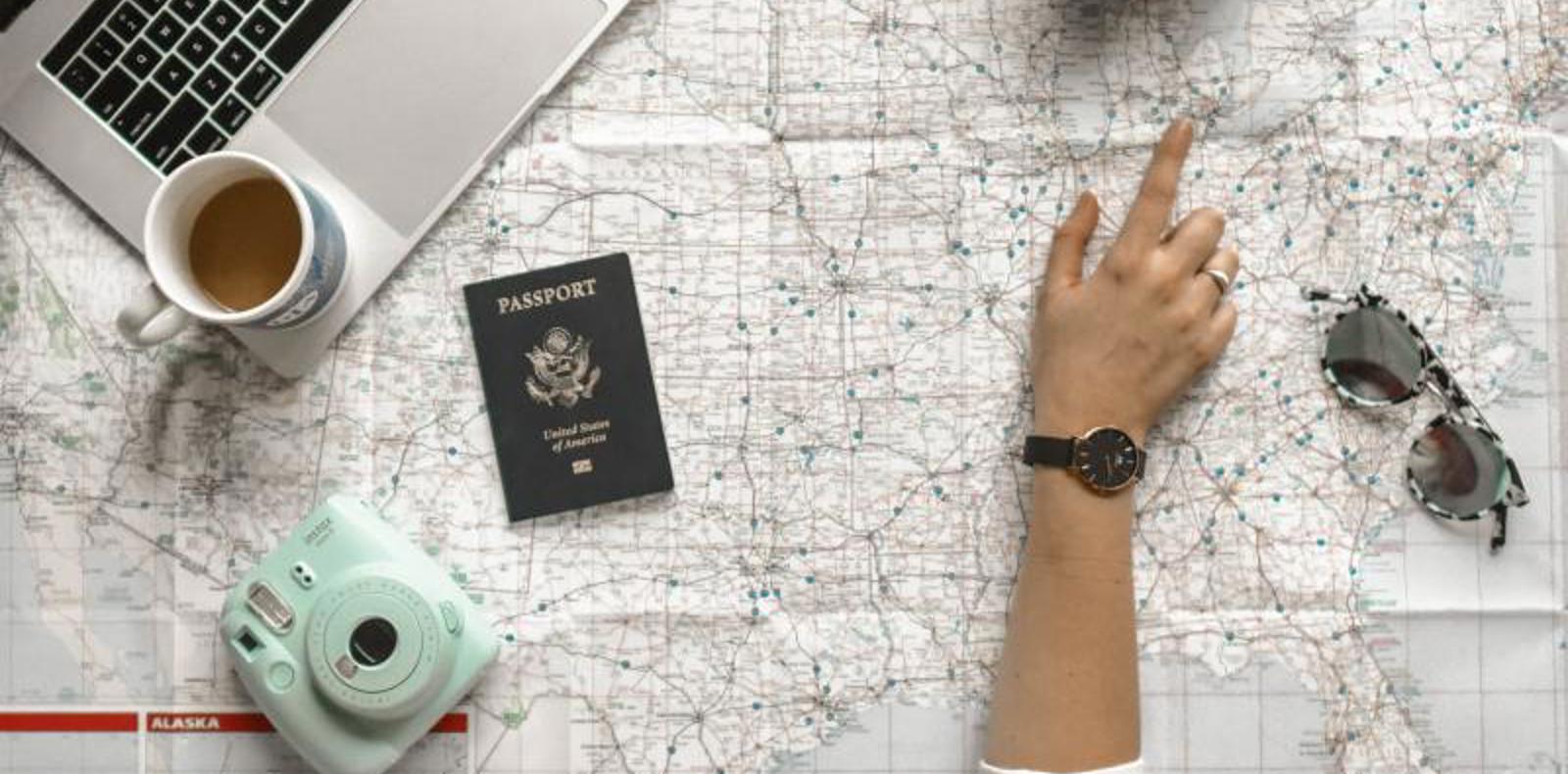 A woman's hand points to a map, laptop, passport, and coffee, indicating travel planning and exploration.