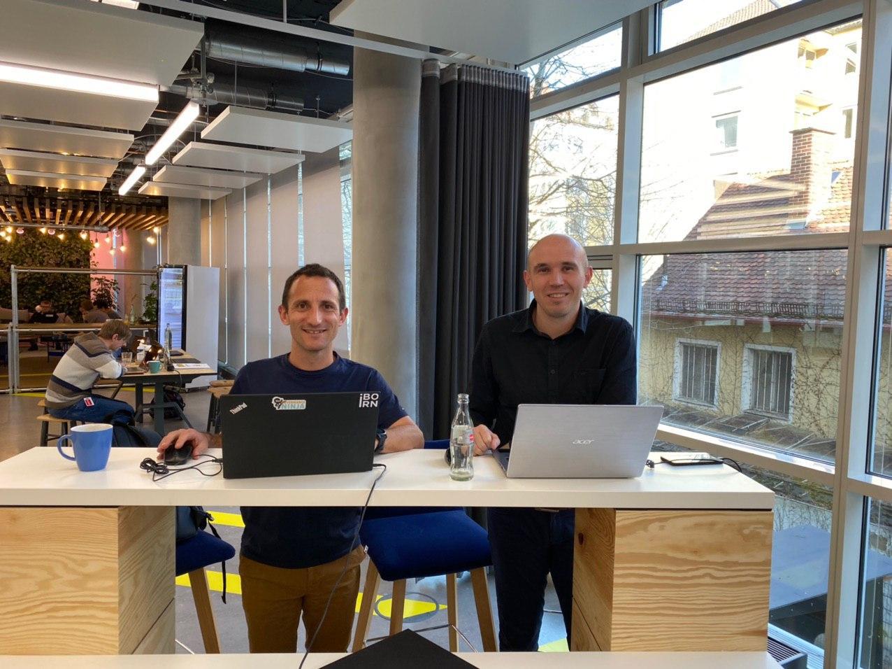 Two men sitting at a desk with laptops on the Amazon RDS Aurora Workshop smiling for a photo. 
