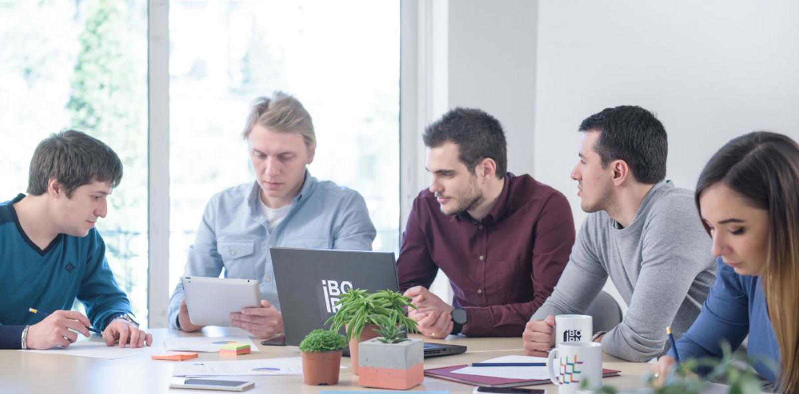 A diverse group of individuals sitting around a table with laptops, engaged in a meeting or collaboration session.